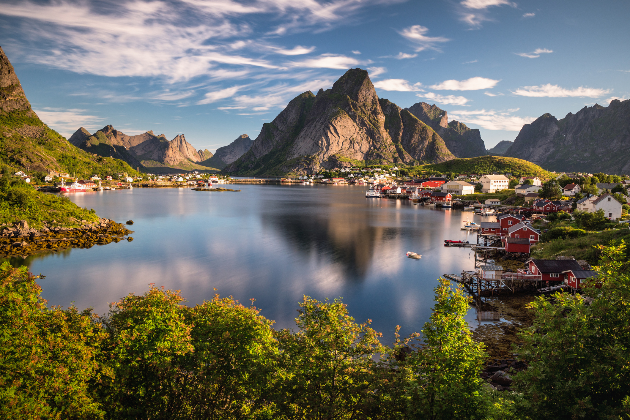 A picturesque waterfront village with houses and boats, set against the backdrop of towering mountains and a clear sky