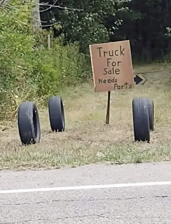 Four tires and a handwritten sign reading "Truck for Sale Needs Parts" are placed on the ground near a road, with an arrow pointing right