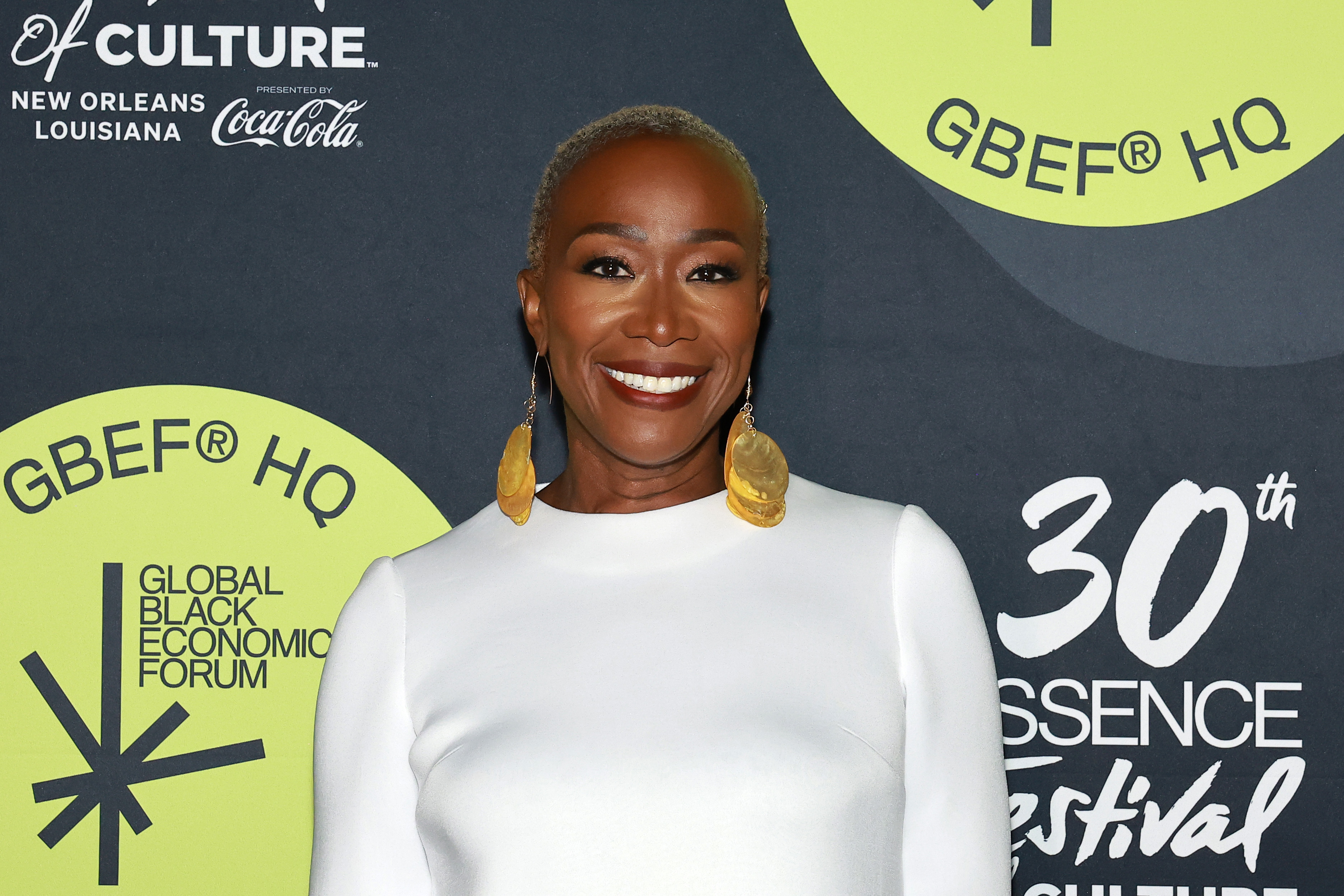 Joy Reid stands smiling in front of a backdrop promoting the 30th Essence Festival of Culture, wearing a sleek, long-sleeved dress and large gold earrings