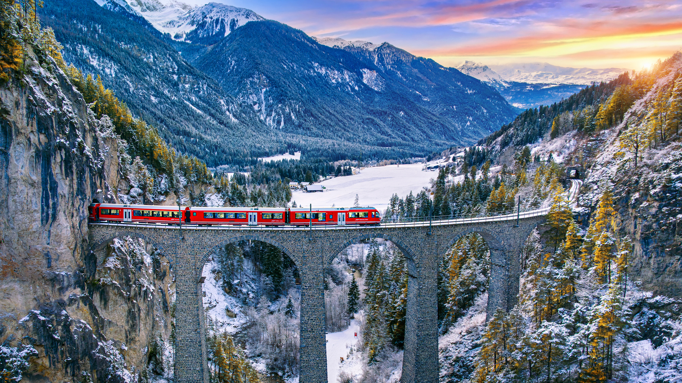 A red train travels across a tall stone bridge surrounded by snowy mountains and a valley at sunrise
