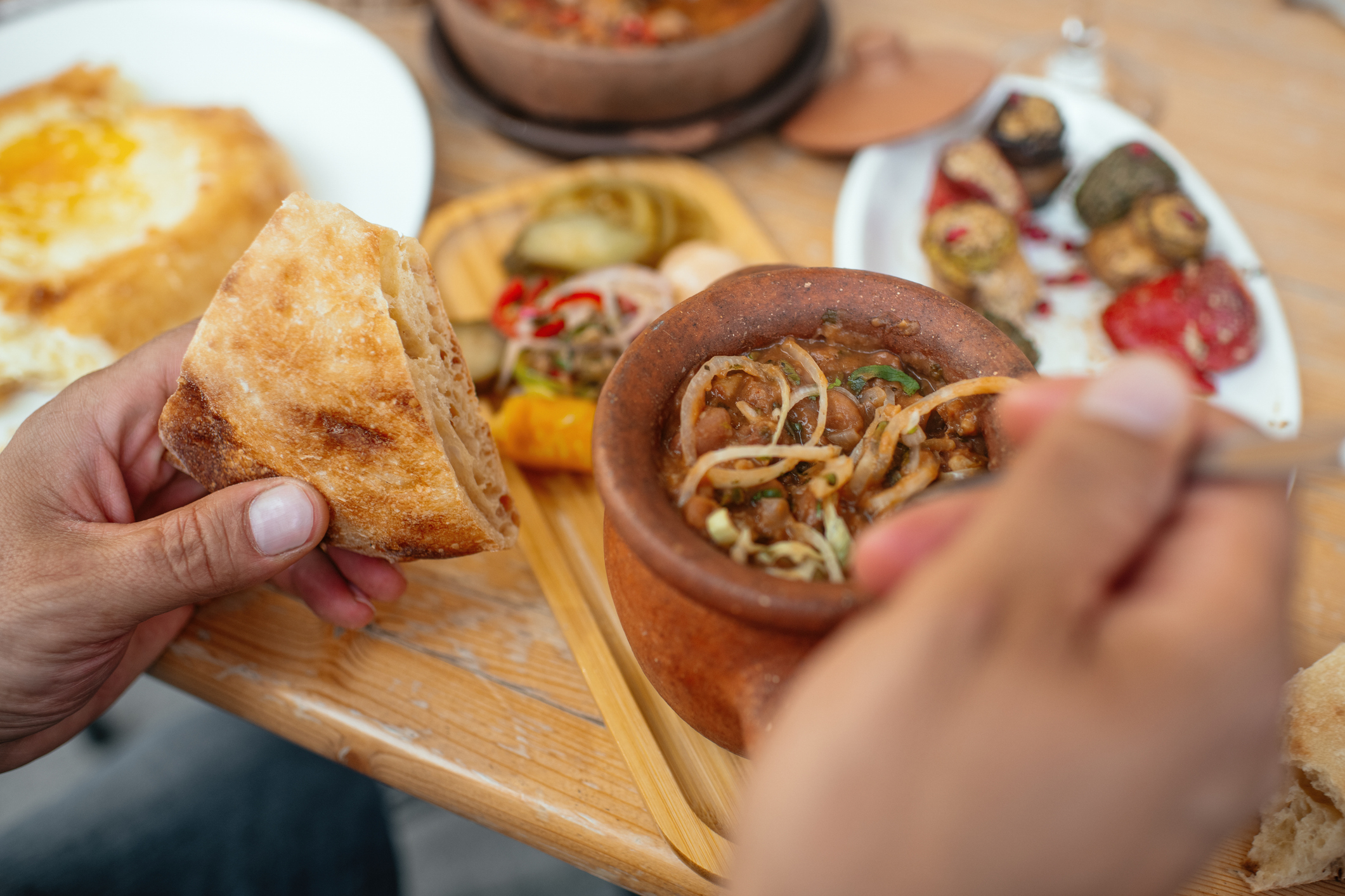 A person is holding a piece of bread and eating a meal consisting of vegetables in a clay pot, with other dishes like pickles and bread on the table