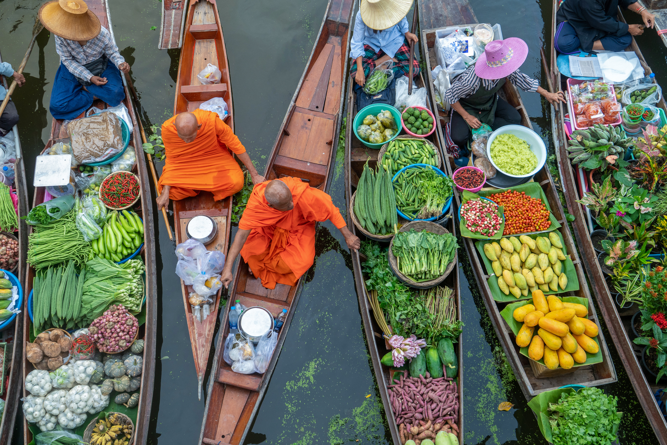 People in boats at a floating market sell various fruits, vegetables, and goods while two monks in orange robes sit in one of the boats laden with packages