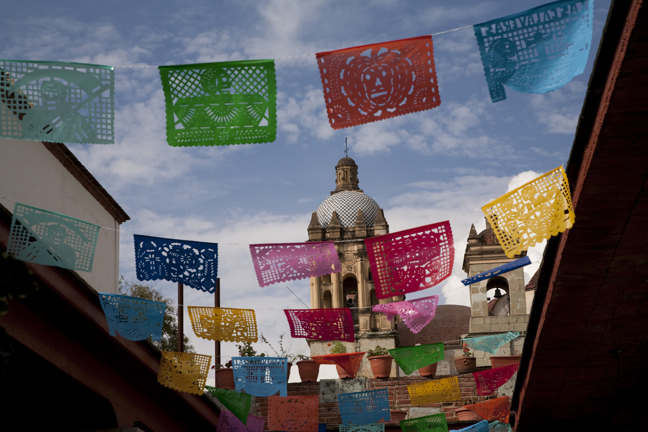 Decorative paper banners, known as papel picado, hang over a street with a domed building and bell tower visible in the background