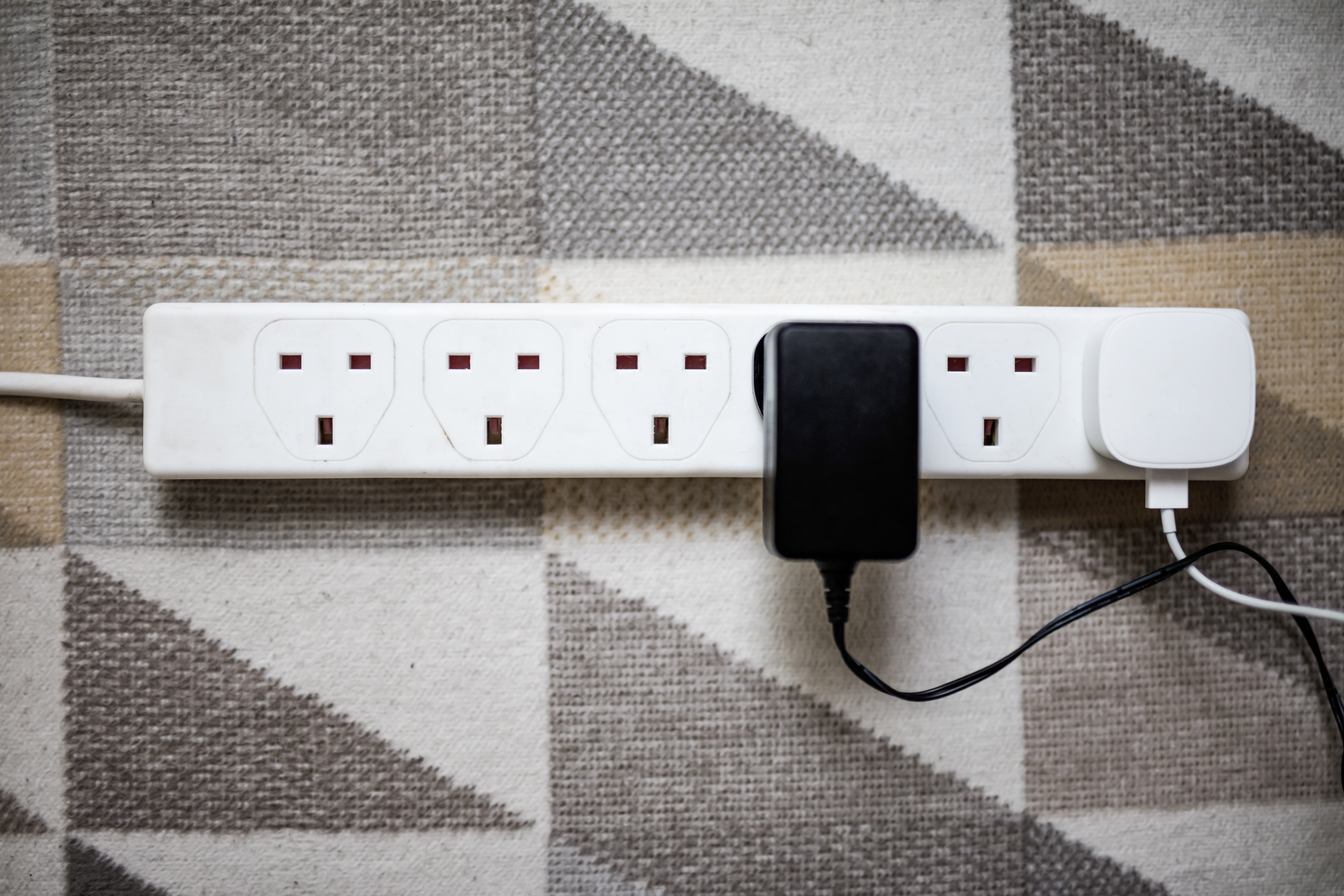 A power strip with four UK sockets is shown, one socket has a black plug, another has a white plug. The strip is set against a geometric patterned surface