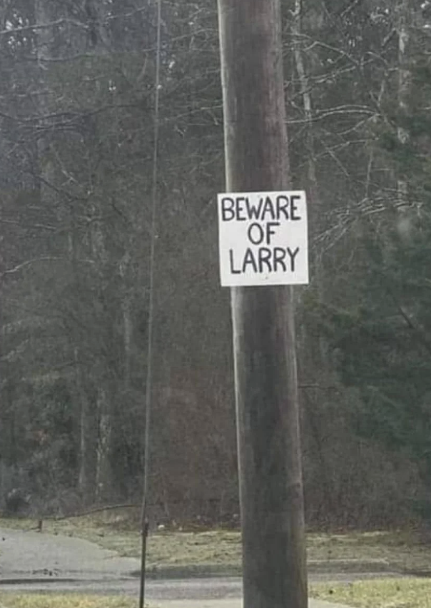 A wooden utility pole with a white sign that reads "Beware of Larry" in hand-drawn capital letters. The background shows a wooded area