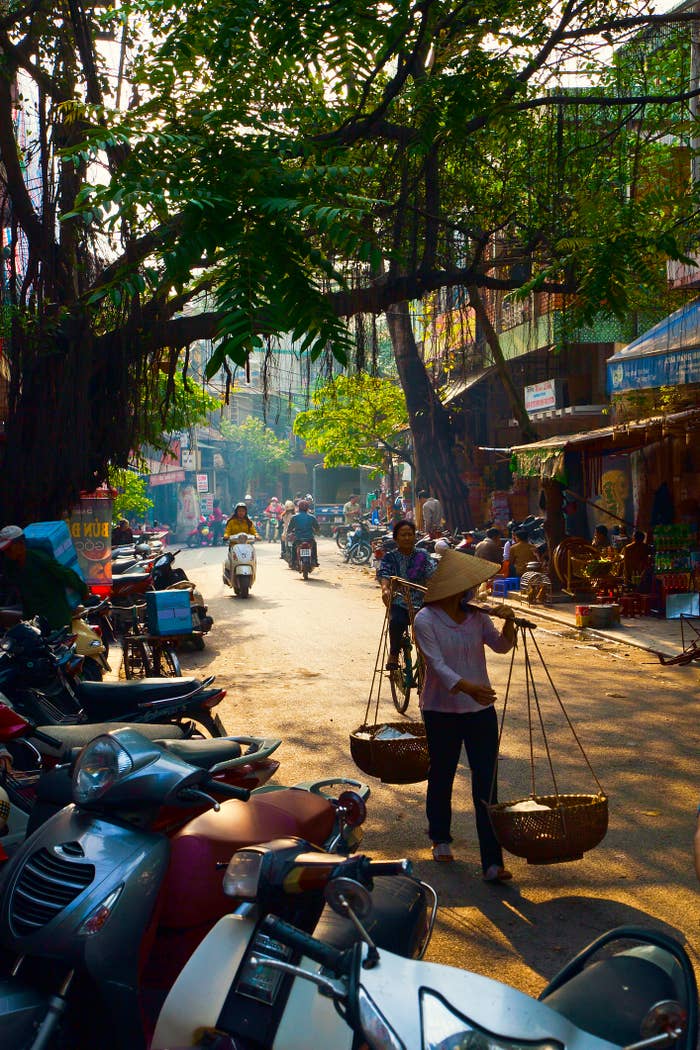Bustling street scene in a vibrant market area with people riding scooters and bicycles. A person walks carrying baskets balanced on a shoulder pole