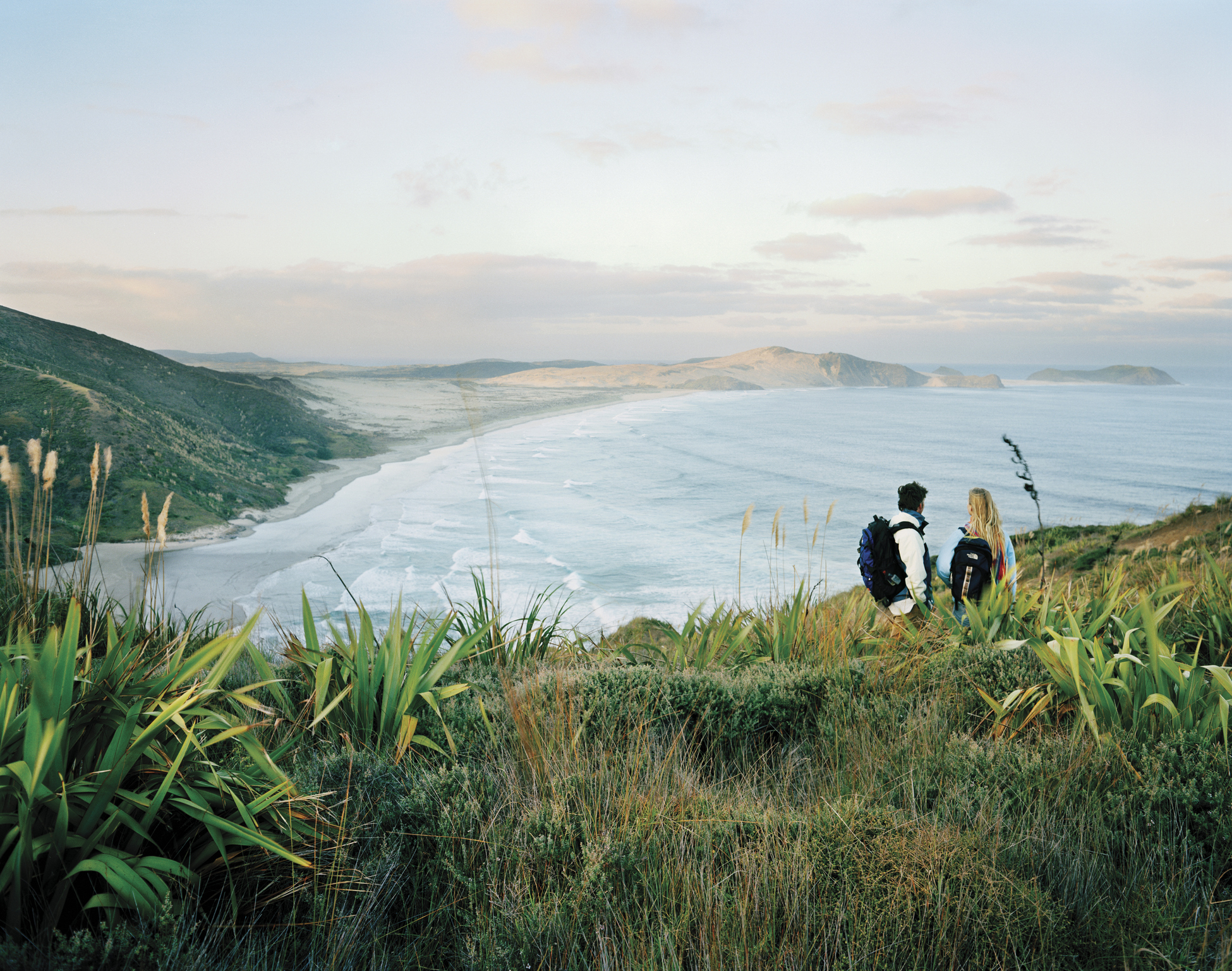 Two hikers with backpacks overlook a vast coastal landscape with waves, beach, and hills in the background