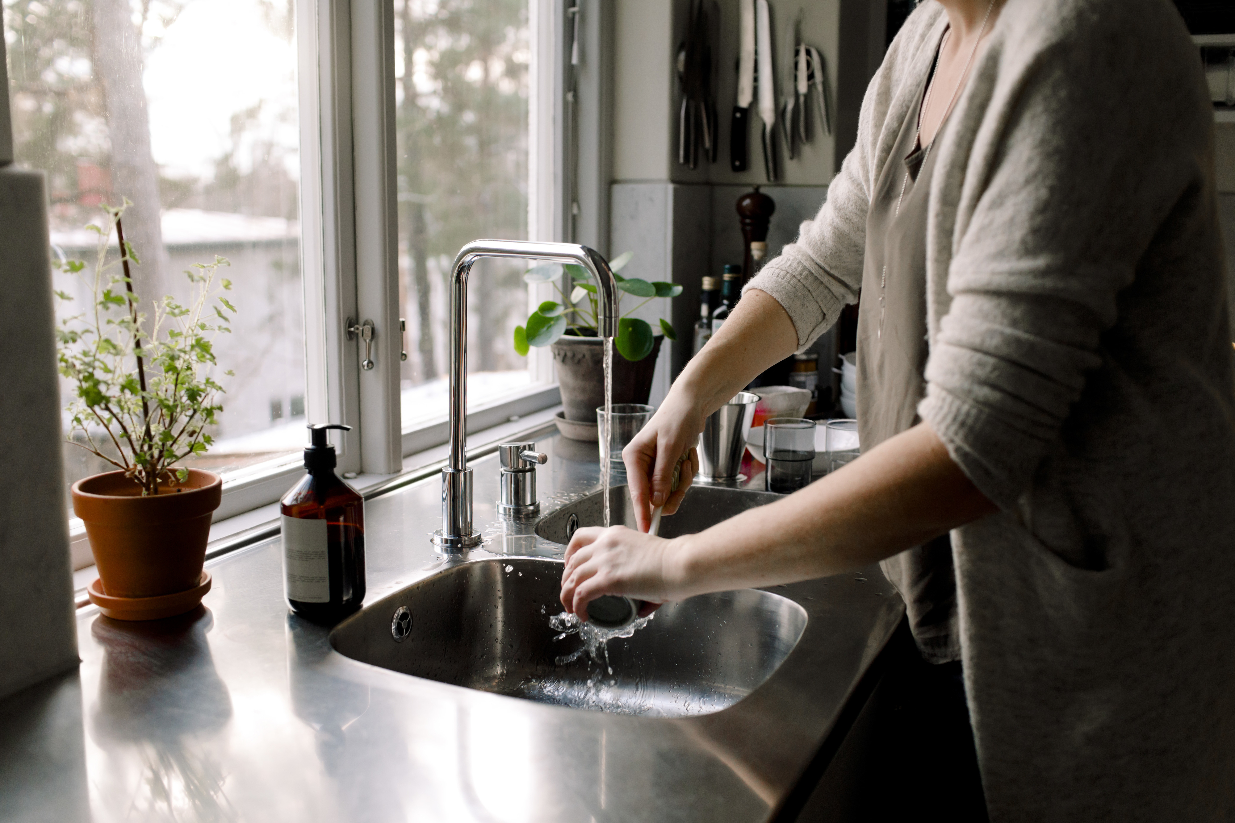 Person washing dishes in a modern kitchen with large windows, potted plants, and a soap dispenser on the counter