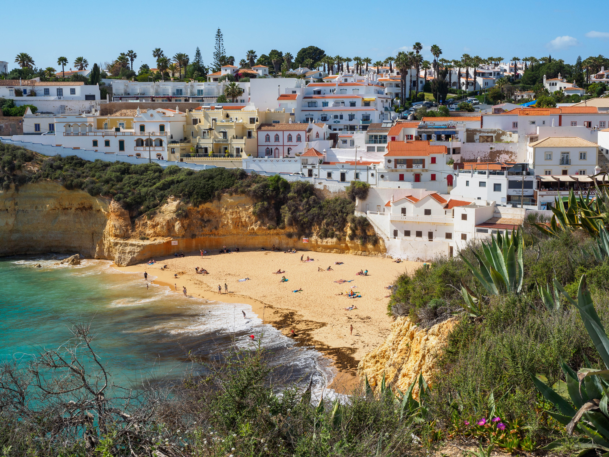 A scenic view of a beach in front of a picturesque coastal town with people relaxing on the sand and swimming in the ocean. Various houses dot the hillside