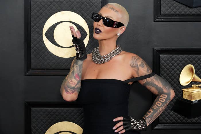 Amber Rose blows a kiss on the red carpet at a celebrity event, dressed in an off-shoulder black gown, large chain necklace, and dark sunglasses, showing her tattoos