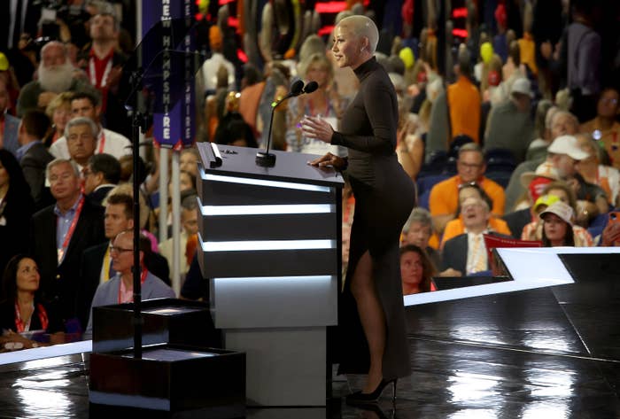 Amber Rose speaks at a podium to a large audience at an event. She wears a form-fitting, long-sleeved dress with a high slit