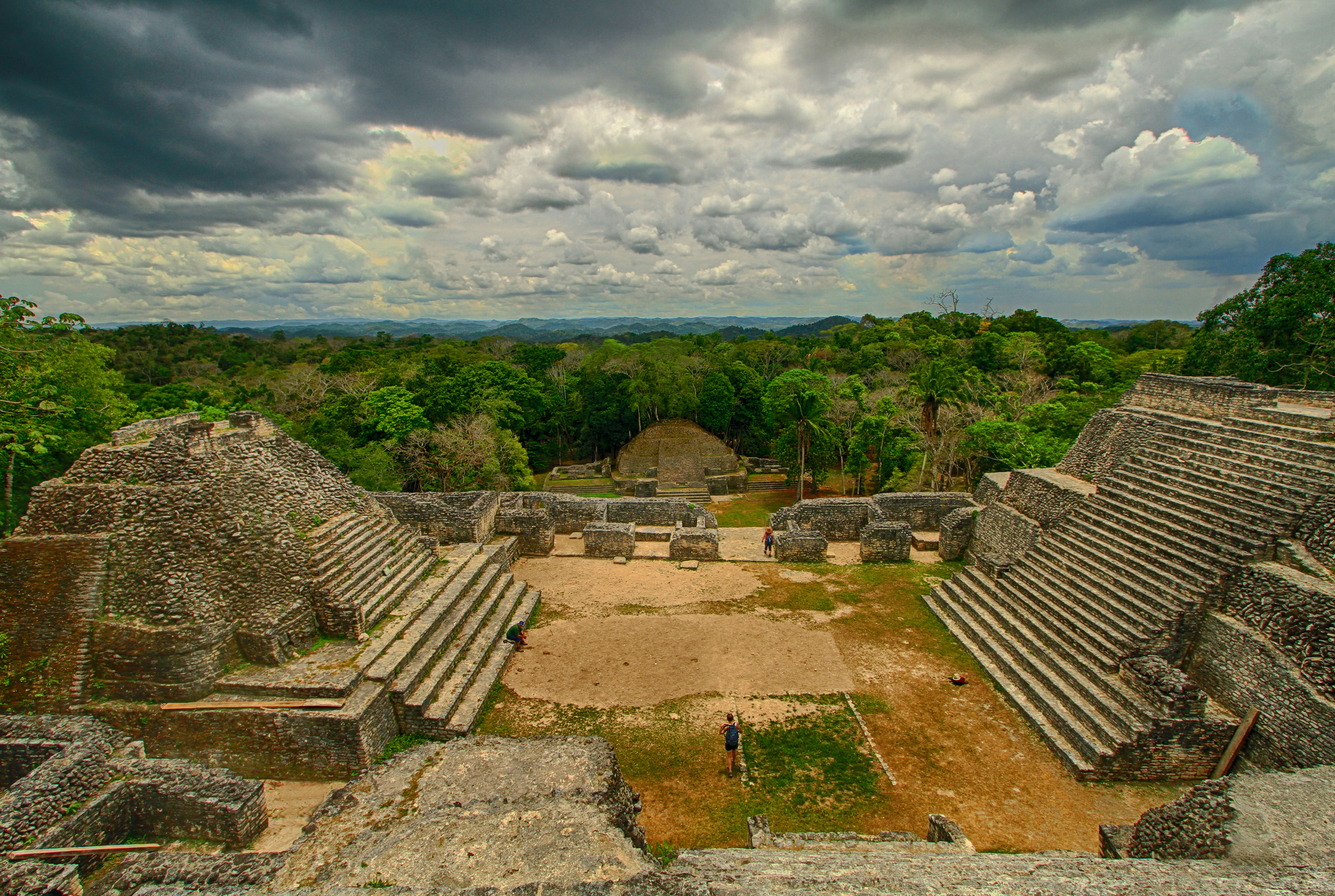 View of ancient Mayan ruins at Caracol in Belize, featuring stone pyramids and staircases with lush jungle in the background under a cloudy sky. No people visible