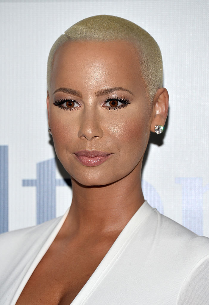 Amber Rose with a short, platinum blonde buzz cut, wearing a V-neck top and stud earrings, poses for a photo at an event