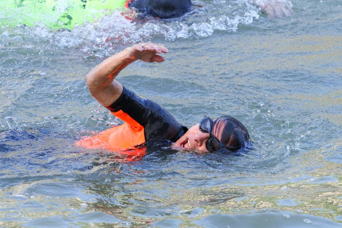 A person wearing a swimsuit, swim cap, and goggles is swimming outdoors in open water with another swimmer visible in the background
