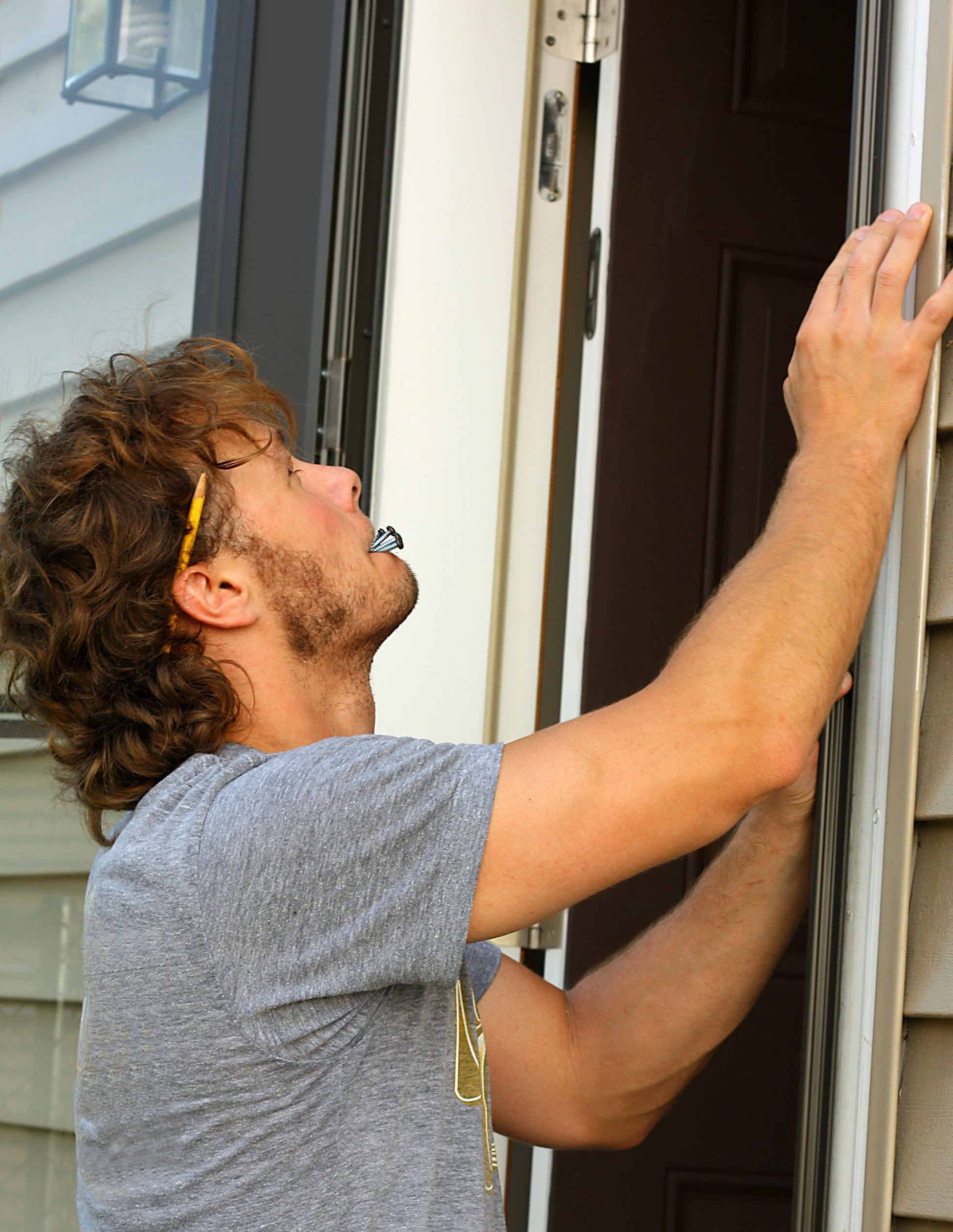 A person in a gray t-shirt looks up while inspecting the siding of a house, with a pencil tucked behind their ear