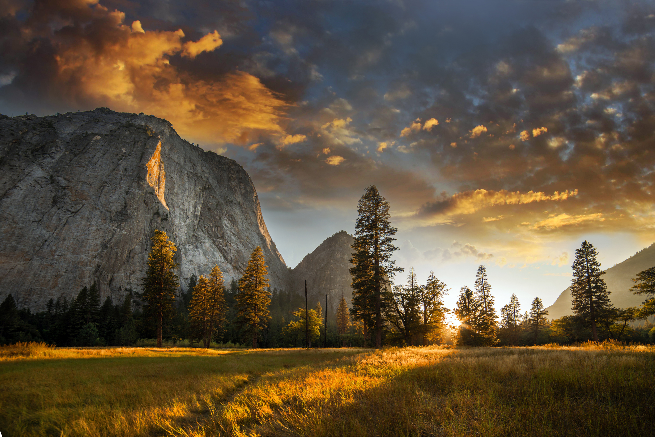Mountain landscape with a dramatic sky, large rock formations, trees, and a sunlit valley
