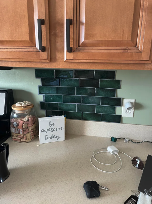 Kitchen counter with jars of dog treats, a motivational sign saying "be awesome today," a cup, car keys, charger, and green tile backsplash