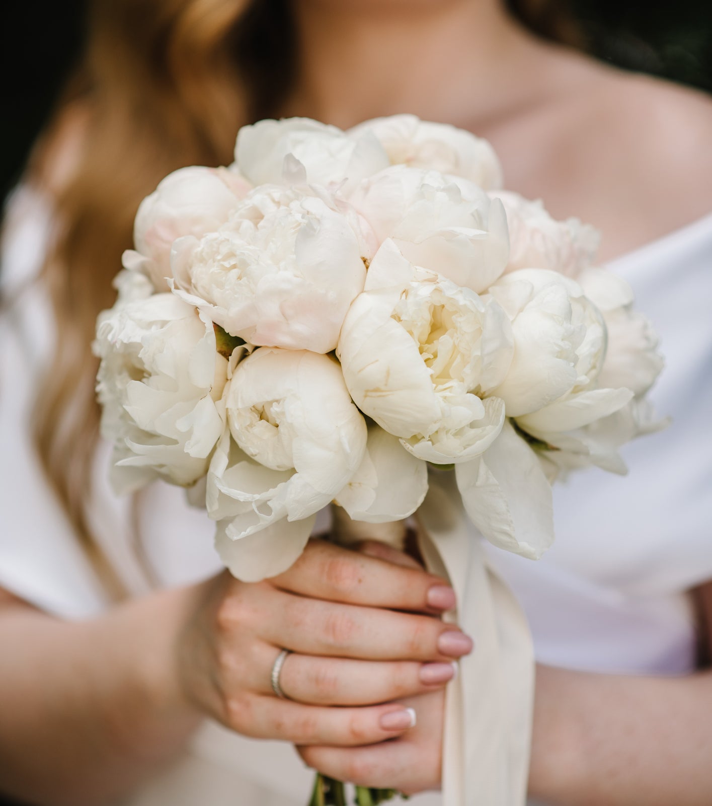 A bride, identity unknown, holds a bouquet of white flowers, wearing an off-the-shoulder dress. Her hair is down, with loose waves
