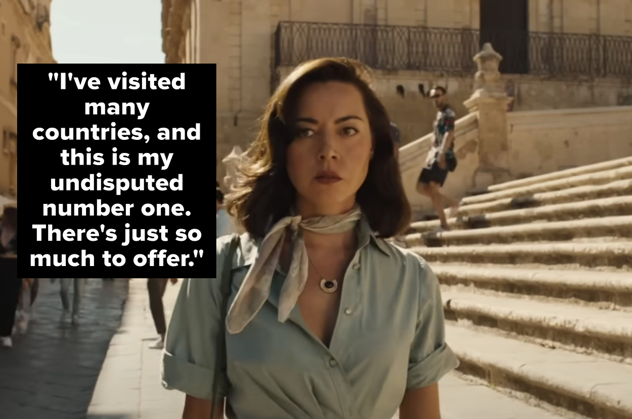 Aubrey Plaza walks through a busy outdoor area with historic stone buildings and steps in the background. She wears a light dress and scarf around her neck