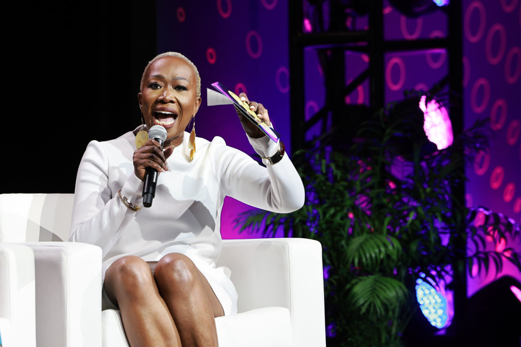 Joy Reid sits on stage holding a microphone and a book, wearing a chic white outfit with statement earrings, speaking passionately at an event