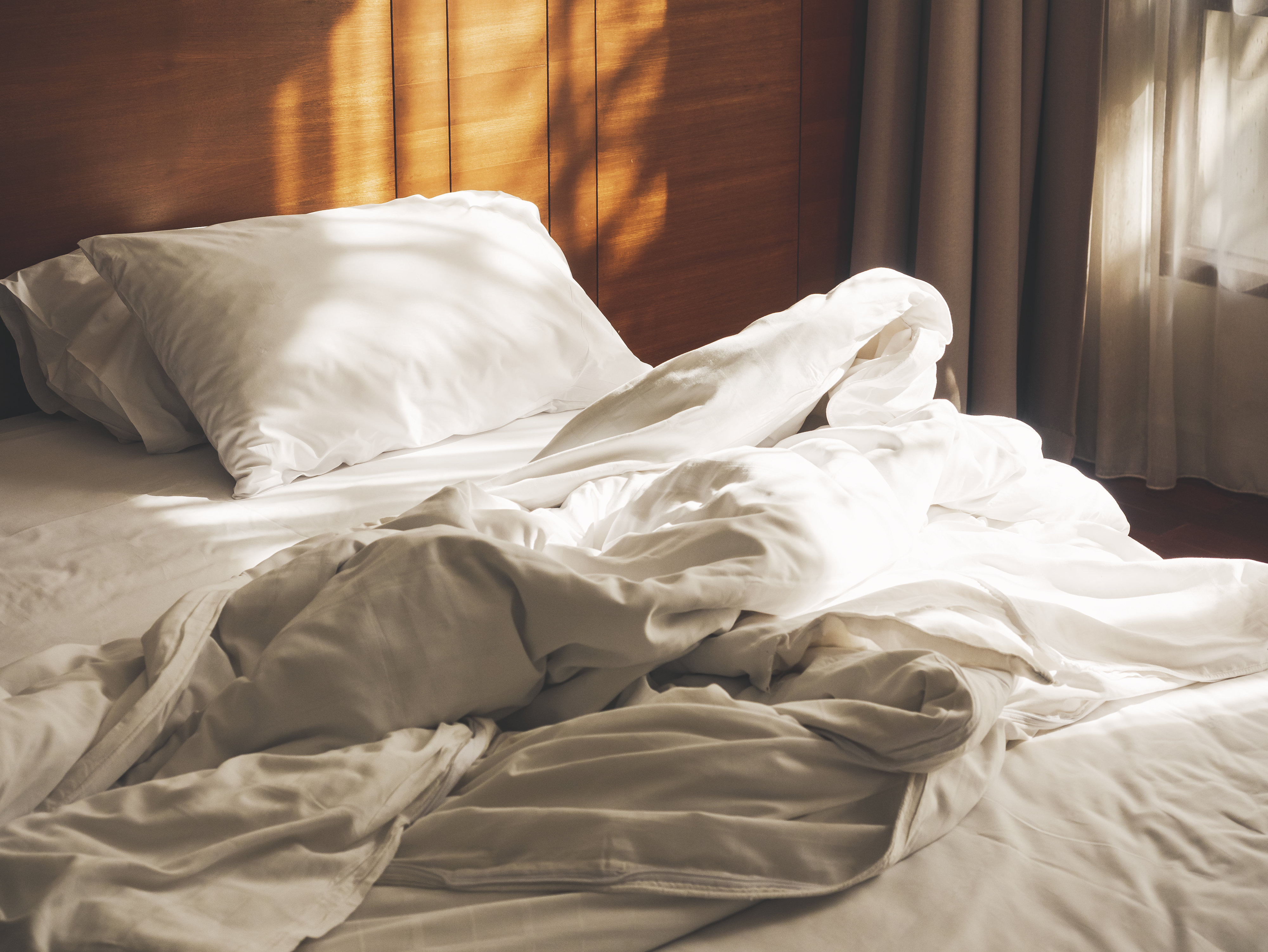 Unmade bed with white sheets and pillows illuminated by sunlight. No people present
