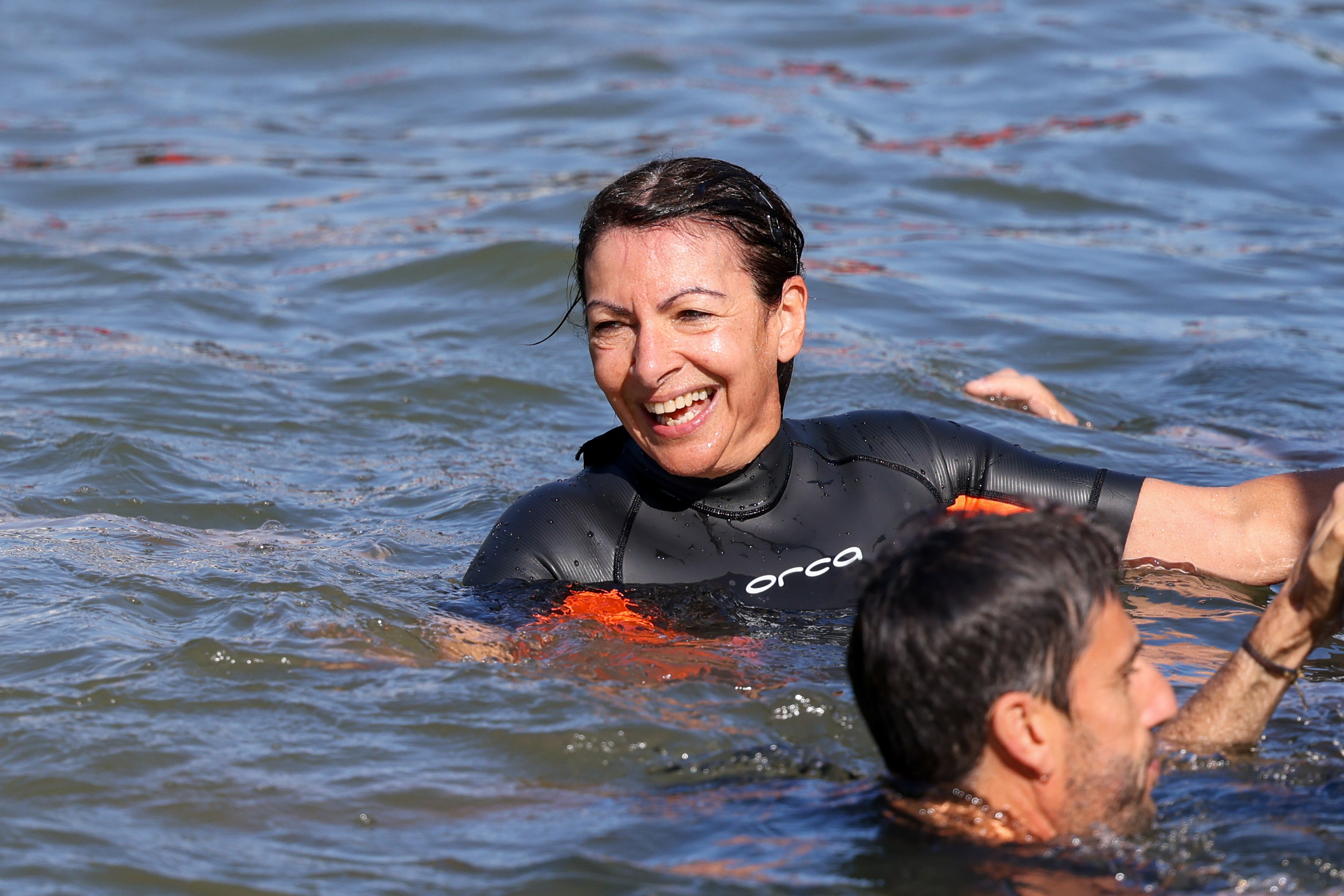 Anne Hidalgo, wearing a wetsuit, smiles while swimming next to a man in open water