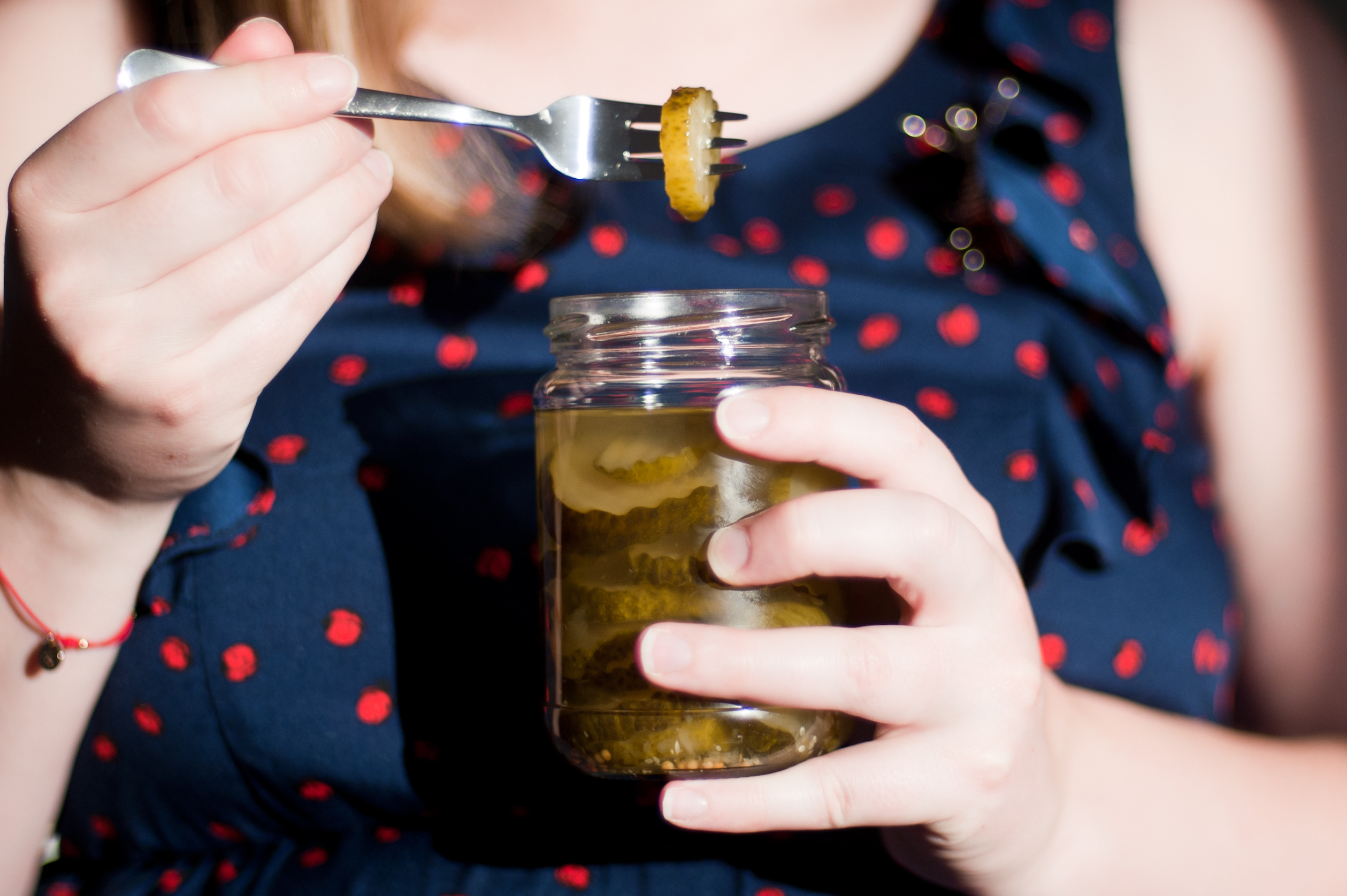 A person in a polka-dot dress holds a jar of pickles with one hand and brings a pickle slice to their mouth with a fork using the other hand