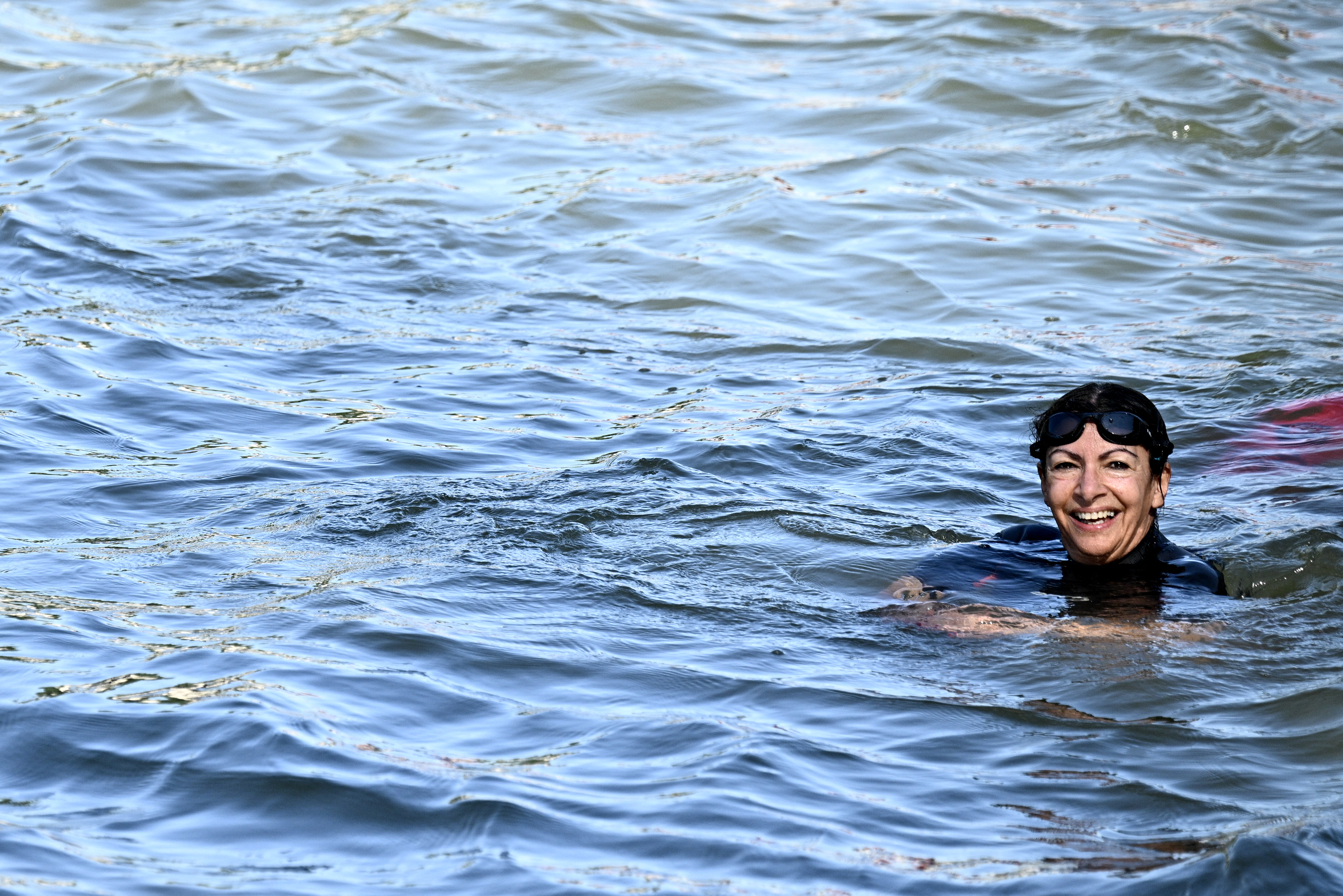 A person wearing swim goggles swims in open water, smiling at the camera