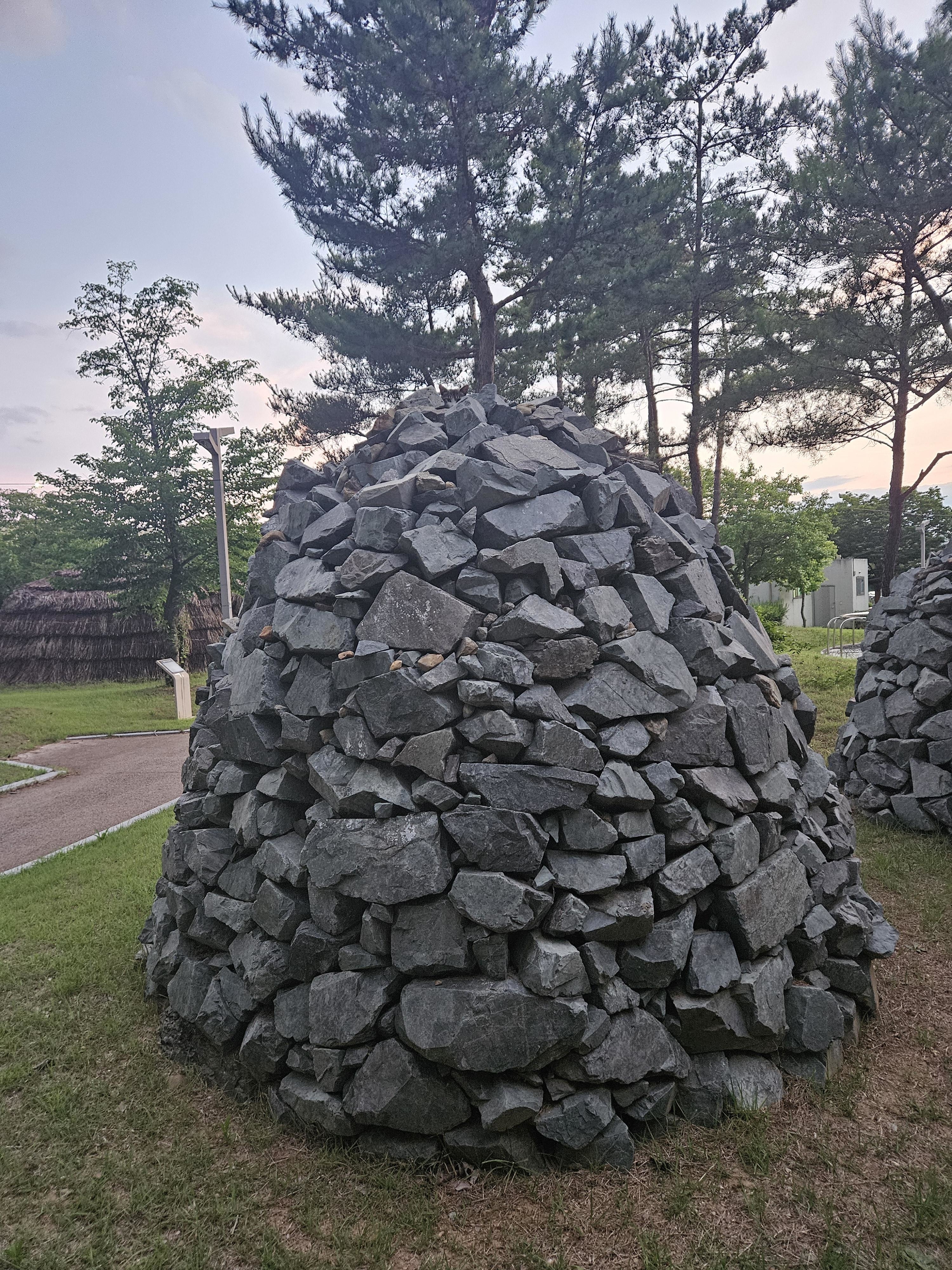 A large conical pile of rocks with a tree growing from the top, set in a park with other trees and pathways in the background