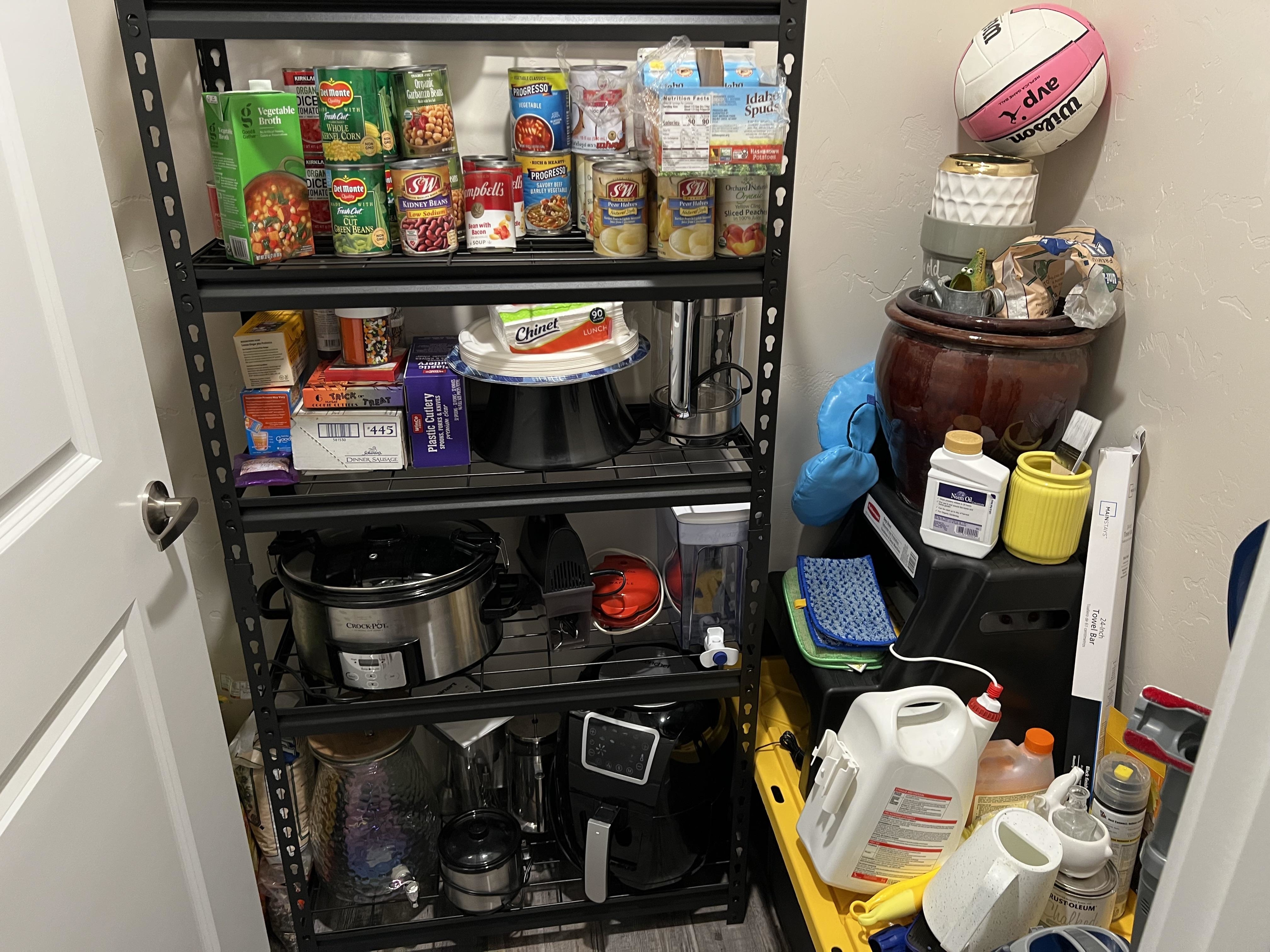 A pantry with shelves containing canned foods, snacks, paper towels, cleaning supplies, kitchen appliances, and a basketball. There are various drinks and cleaners on the floor