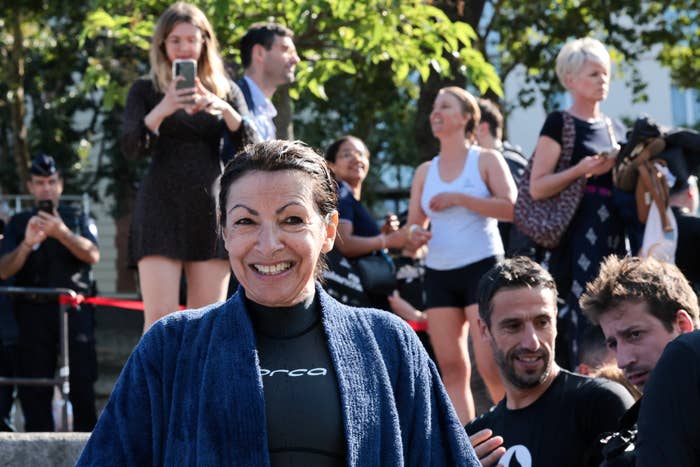 A woman in a wetsuit and robe smiles at the camera with a group of people in the background, some taking photos