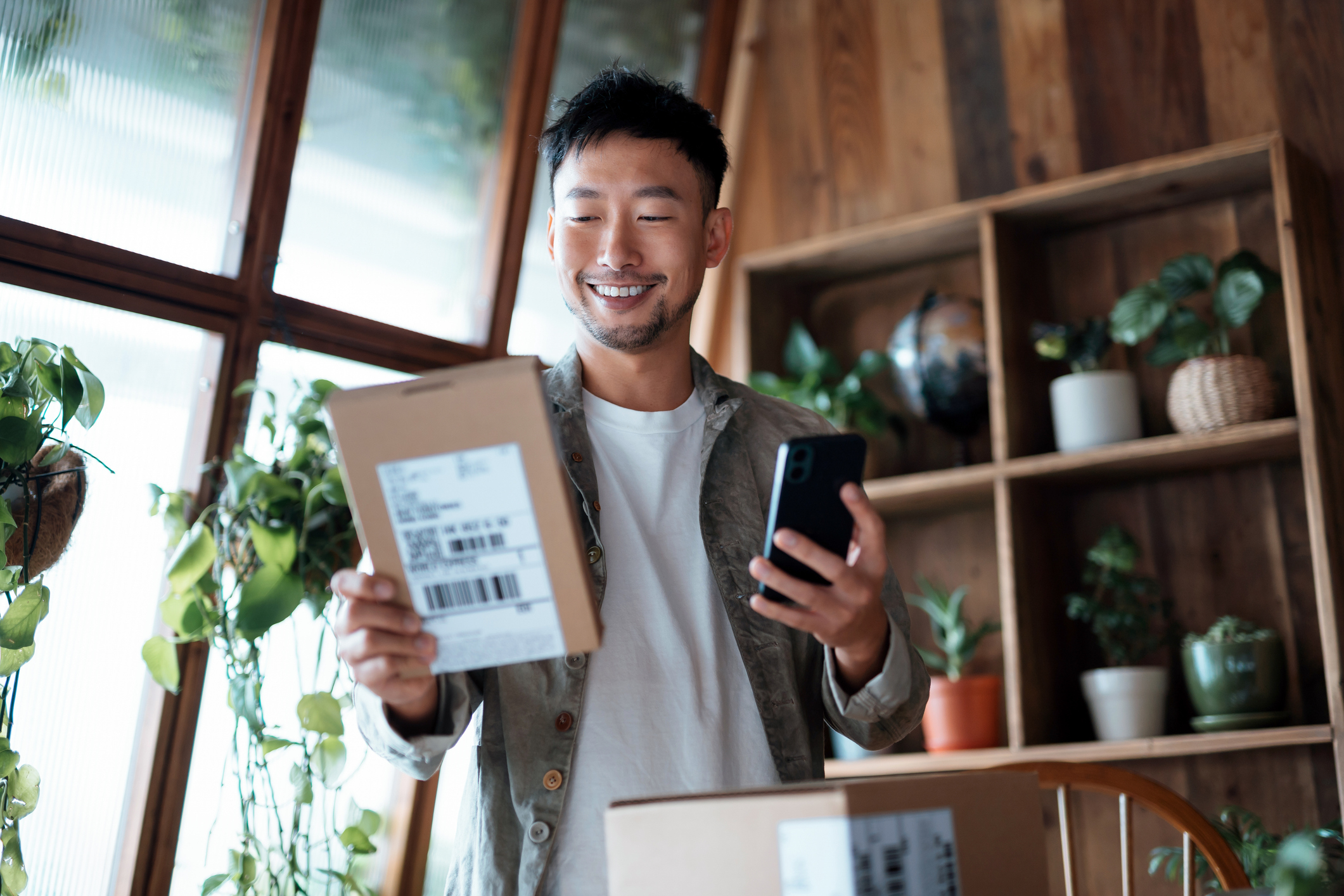 A person stands smiling while holding a package and a phone, in a room filled with plants and wooden decor in the background