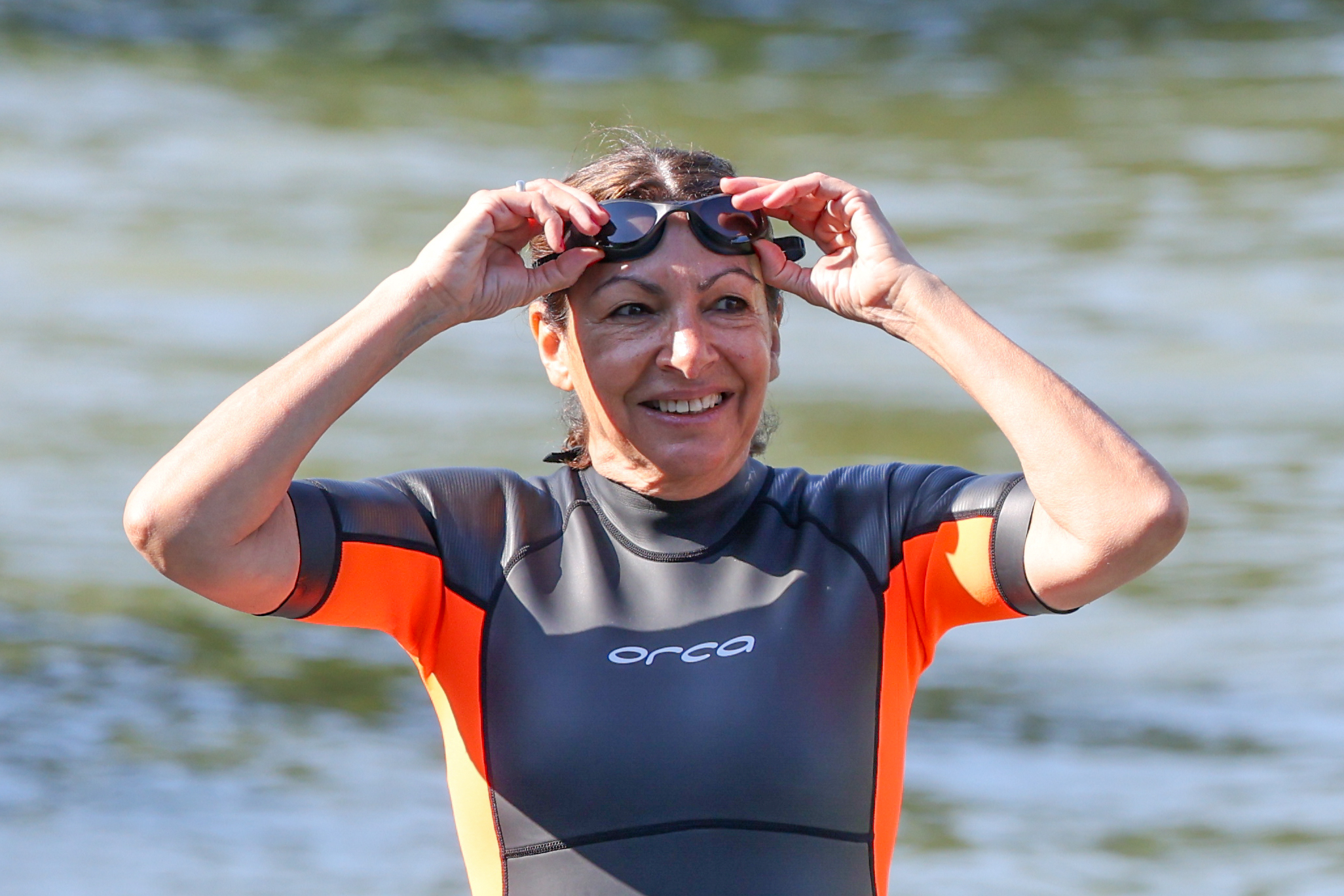 A woman outstretched arms above her head pulling her goggles over her eyes wearing a wetsuit with "orca" on the chest, standing in front of a body of water