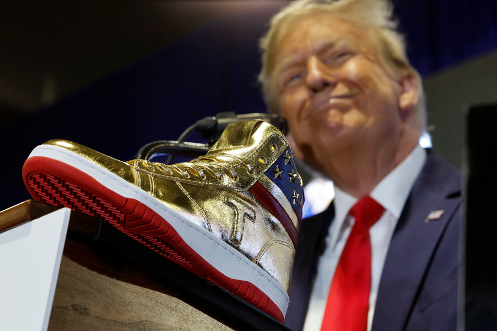 Donald Trump smiling behind a podium with a gold sneaker displayed on it. The sneaker features a star design
