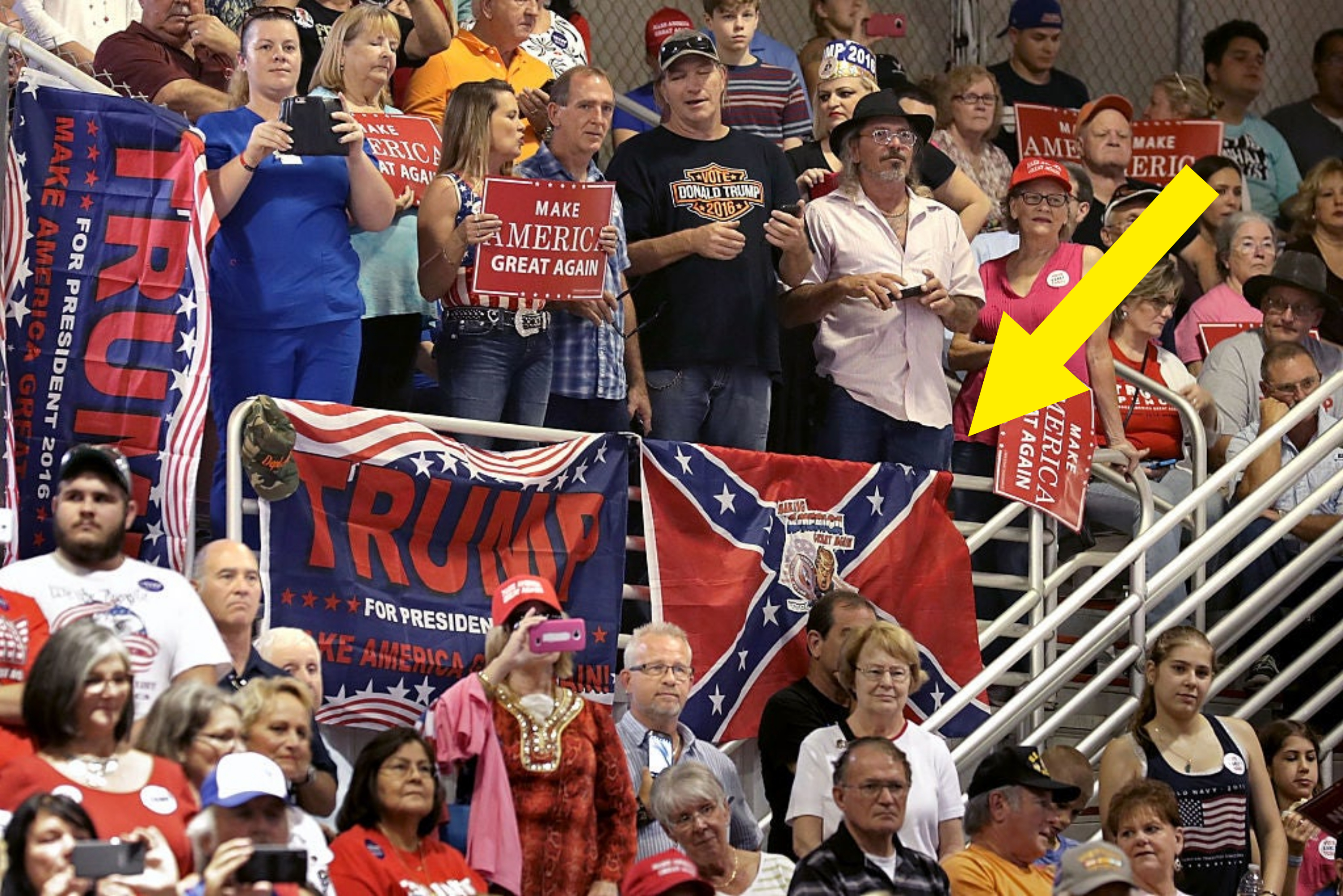 A crowd of people holding "Make America Great Again" and "Trump" signs at a political rally. A Confederate flag is hanging among several Trump campaign banners