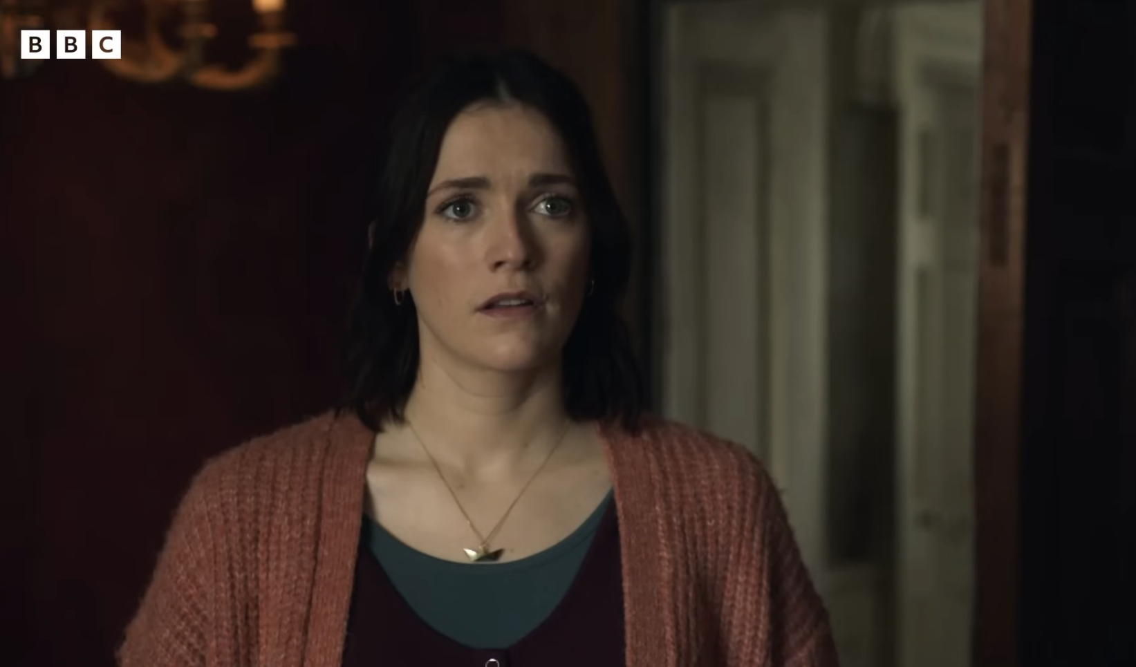 A woman with dark, shoulder-length hair, wearing a cardigan and necklace, looks concerned in a scene from a BBC show
