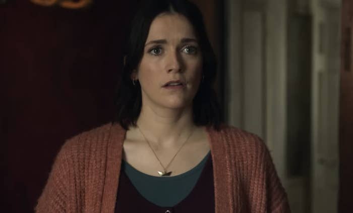 A woman with dark, shoulder-length hair, wearing a cardigan and necklace, looks concerned in a scene from a BBC show