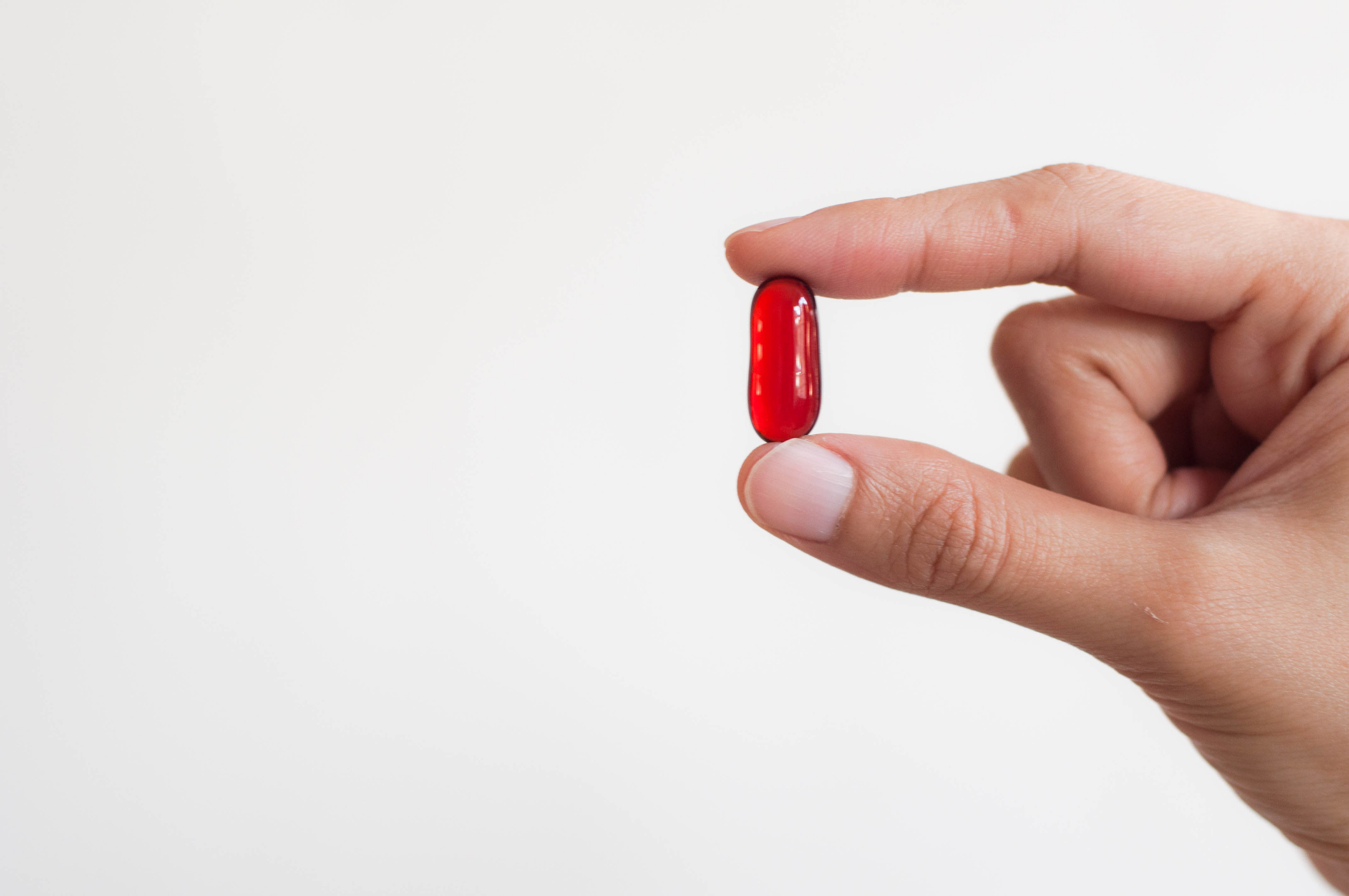 A hand holding a single red capsule pill between the thumb and index finger against a plain background