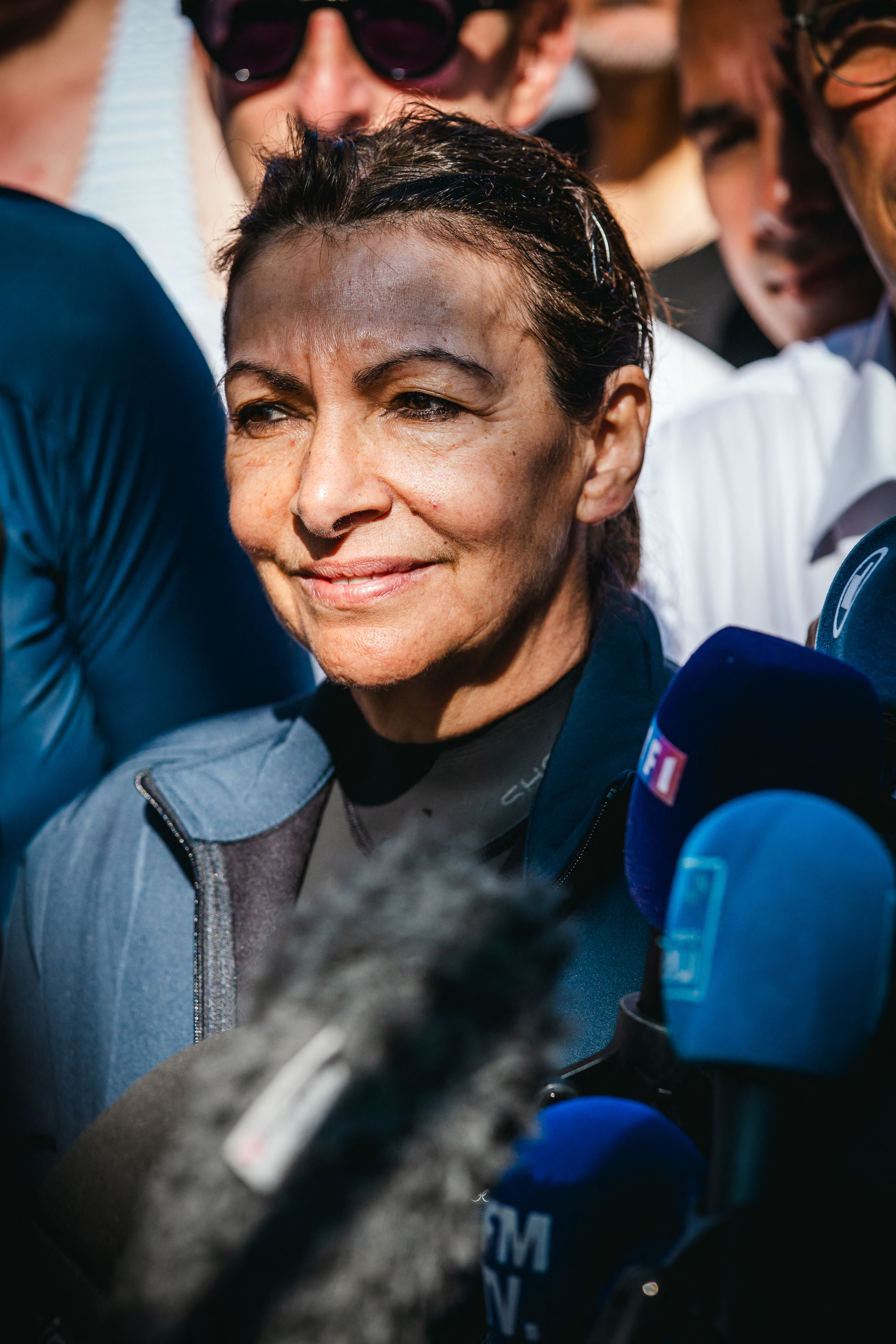 Anne Hidalgo stands among reporters and microphones, smiling slightly. She wears a sports jacket and appears to be at an outdoor event