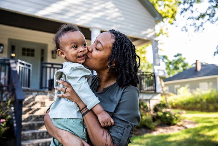 A person holding and kissing a smiling baby on the cheek, standing in front of a house