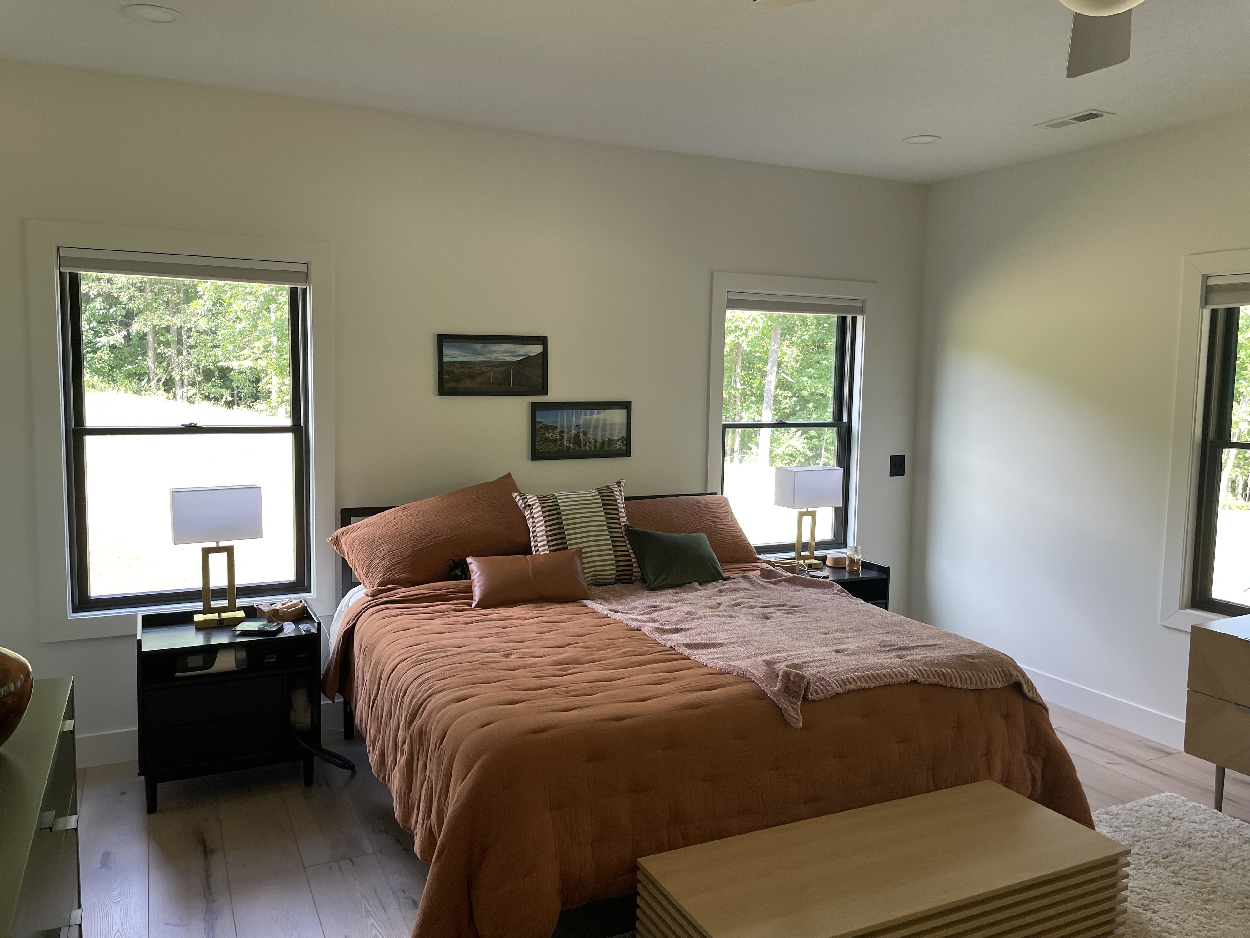 A neatly made bed with pillows and a throw blanket in a minimalistic bedroom. Two windows with scenic views flank the bed, and bedside tables hold lamps
