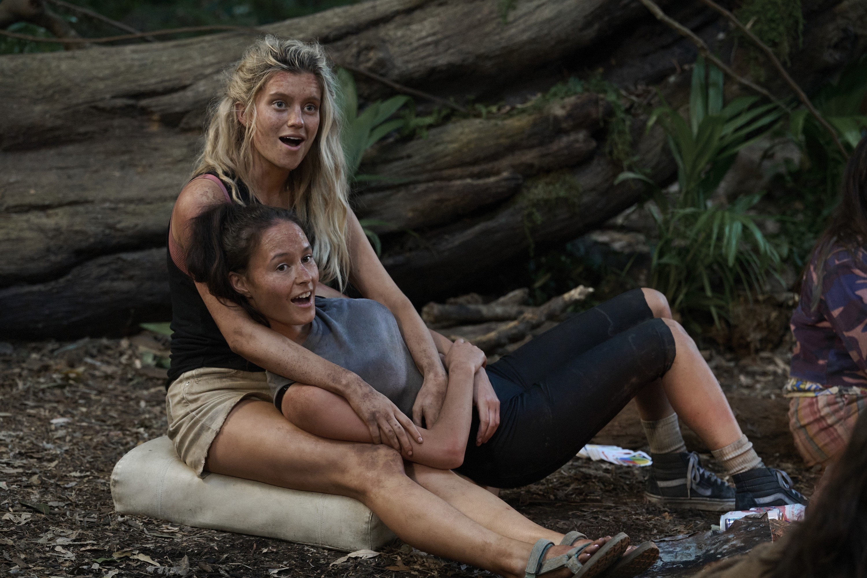 Erana James, wearing a casual shirt and shorts, sits on the ground leaning against Mia Healey, who is embracing her. Both appear to be in a forest setting