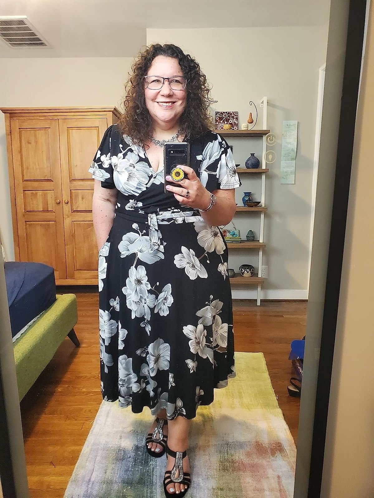 A smiling reviewer with curly hairsbreadth  wears a floral-patterned formal  and sandals portion    taking a reflector  selfie successful  a well-decorated room