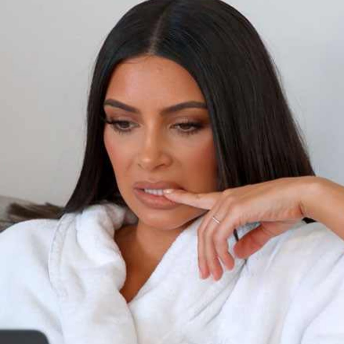 Kim Kardashian wearing a white robe, looking at something off-camera with a thoughtful expression and one finger on her lips