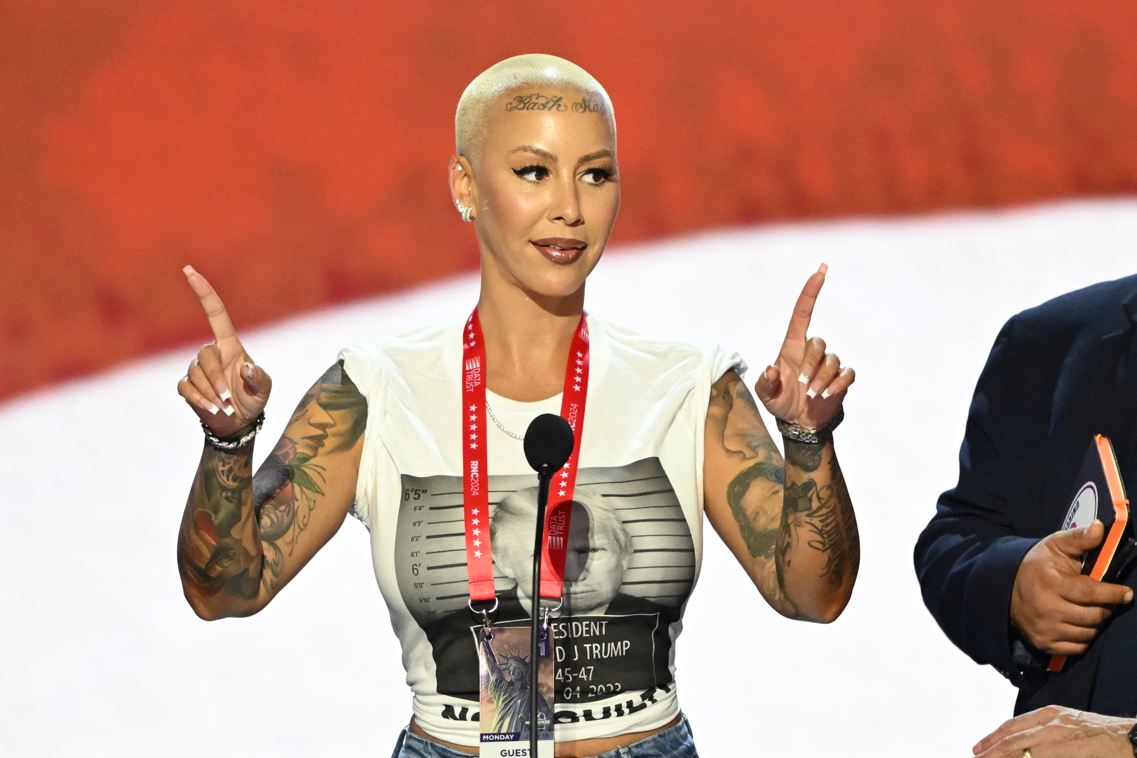 Amber Rose speaking at an event, wearing a t-shirt with Donald Trump's mugshot print, pointing upwards with both hands. Microphone and lanyard present