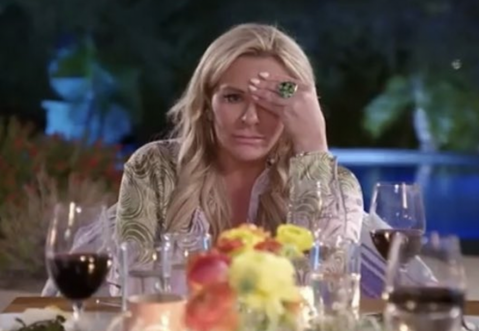 Woman with long hair sits at an outdoor dinner table, resting face on one hand, surrounded by wine glasses and flowers