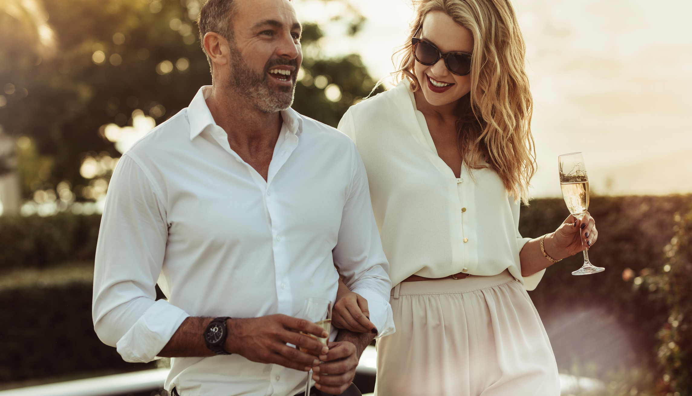 A man and a woman smile and walk outdoors. The man wears a white shirt and the woman, in sunglasses, wears a white blouse and light trousers, holding champagne
