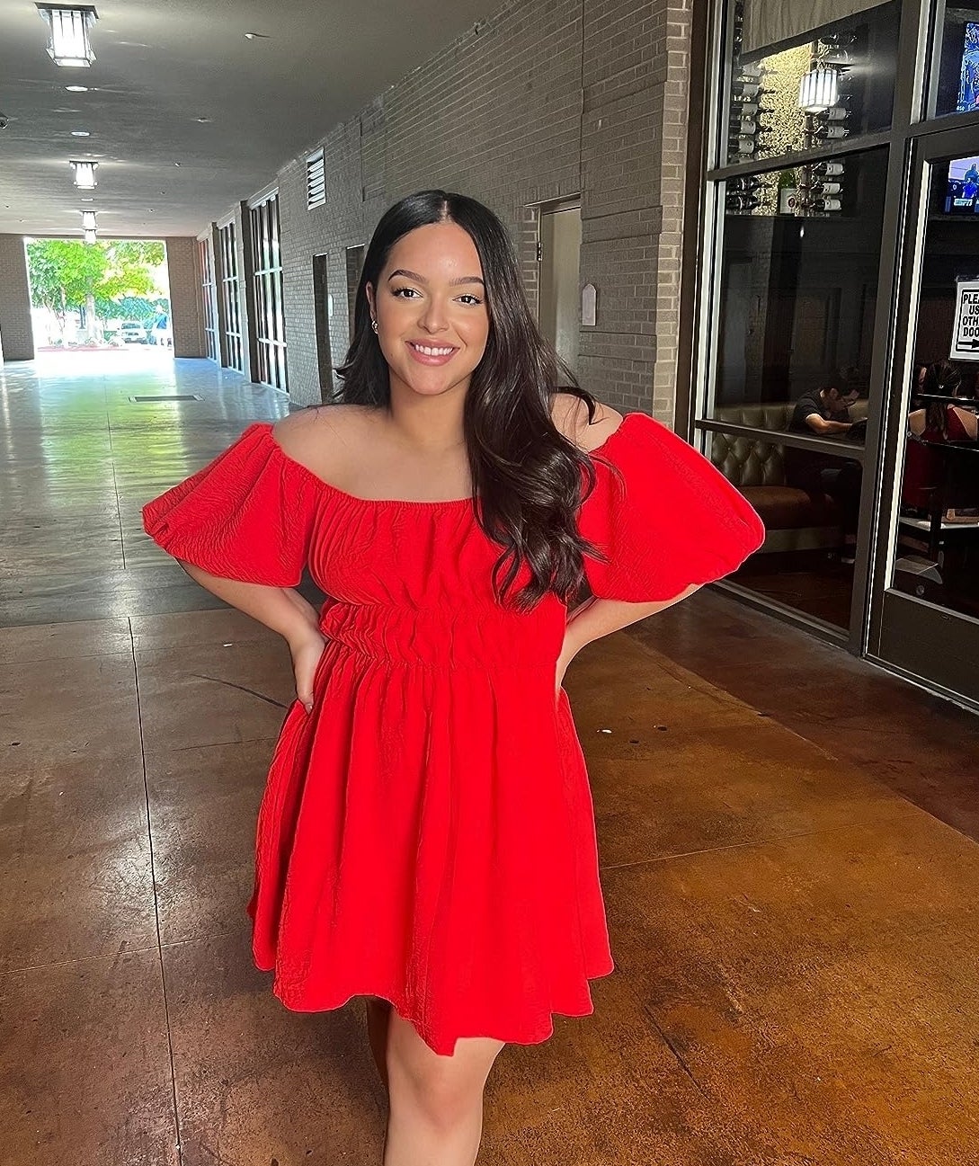A reviewer stands in a hallway wearing an off-the-shoulder red dress, smiling with hands on hips