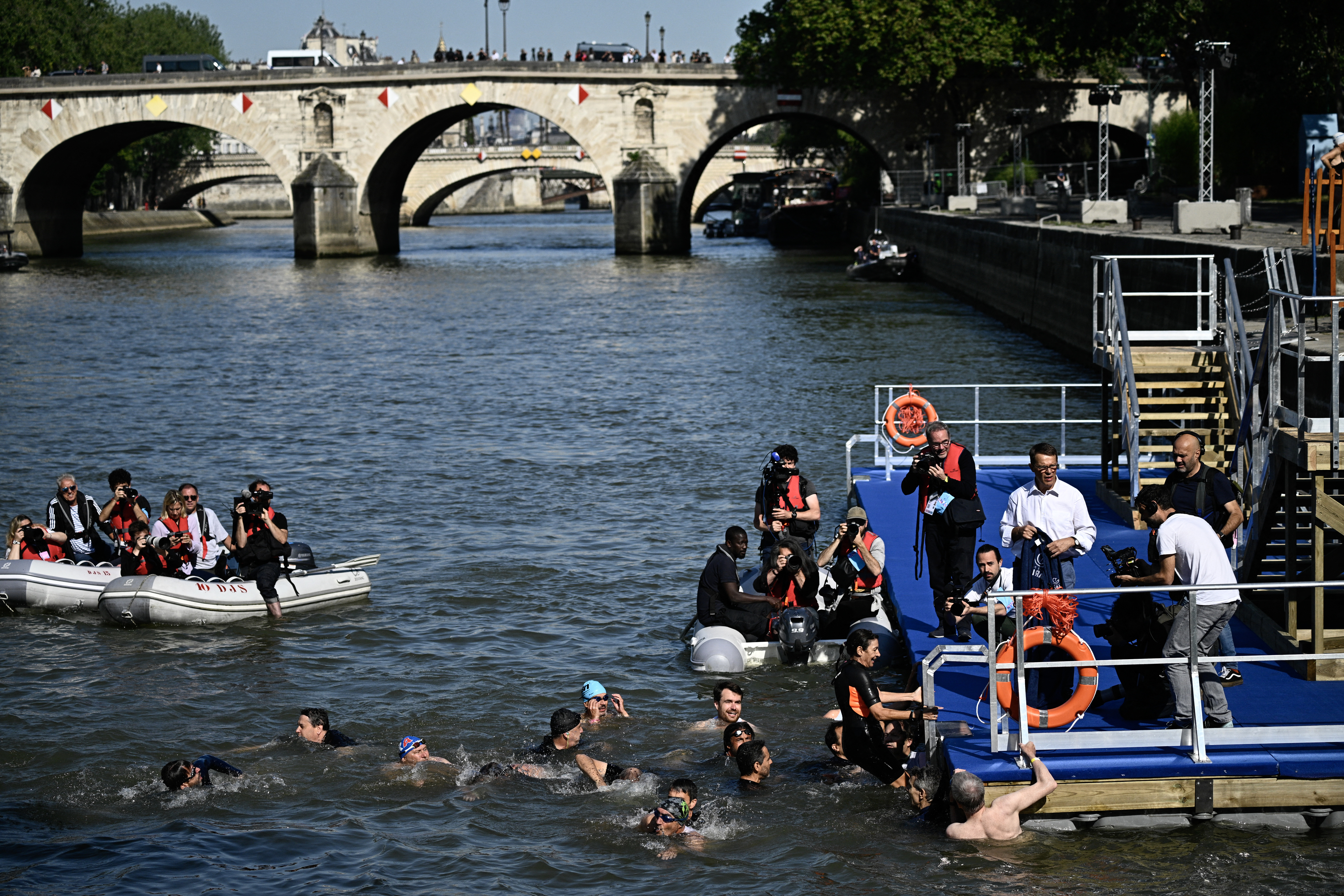 Participants swim in the Seine River under a bridge during an event, with some climbing out onto a dock, surrounded by safety staff and boats