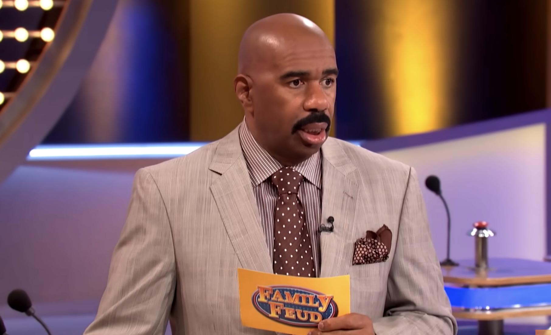 Steve Harvey hosts &quot;Family Feud,&quot; wearing a light-colored suit and polka-dot tie while holding a game card. The set and buzzer podiums are visible behind him
